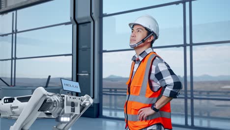 vista lateral de un ingeniero masculino asiático con casco de seguridad en una fábrica de alta tecnología, analizando el concepto de la máquina robótica. de pie con los brazos akimbo mirando a su alrededor, comprobando
