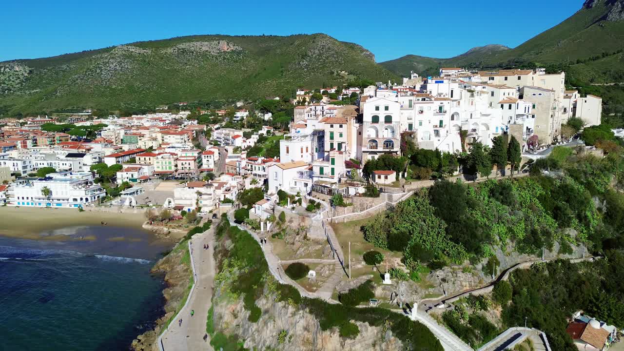 ascenso aéreo de la costa soleada de sperlonga, lazio, con edificios encalados y encanto mediterráneo mientras los turistas caminan debajo