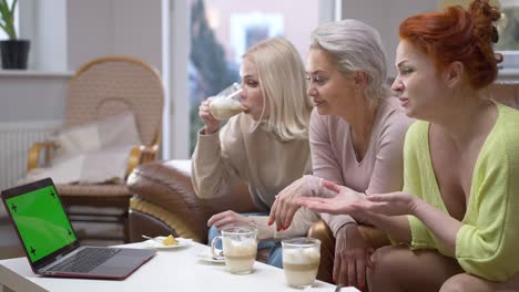 three positive women sitting on couch waving at laptop with chromakey green screen smiling. happy caucasian friends greeting mockup video chat resting on weekend together indoors