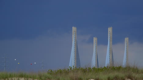 Zeitrafferaufnahme-Einer-Brücke-Und-Stromleitungen-Mit-Dünen-Im-Vordergrund-Und-Dramatischen-Wolken-Am-Himmel