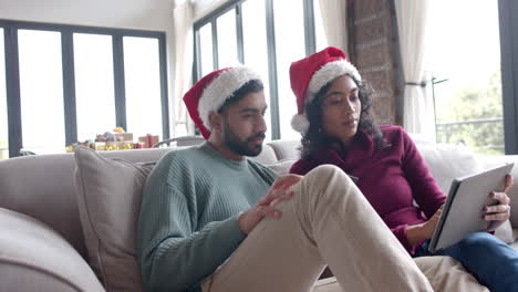 Pareja-Birracial-Con-Sombreros-De-Santa-Claus-Sentados-En-Un-Sofá-Usando-Una-Tableta-En-Casa,-Cámara-Lenta