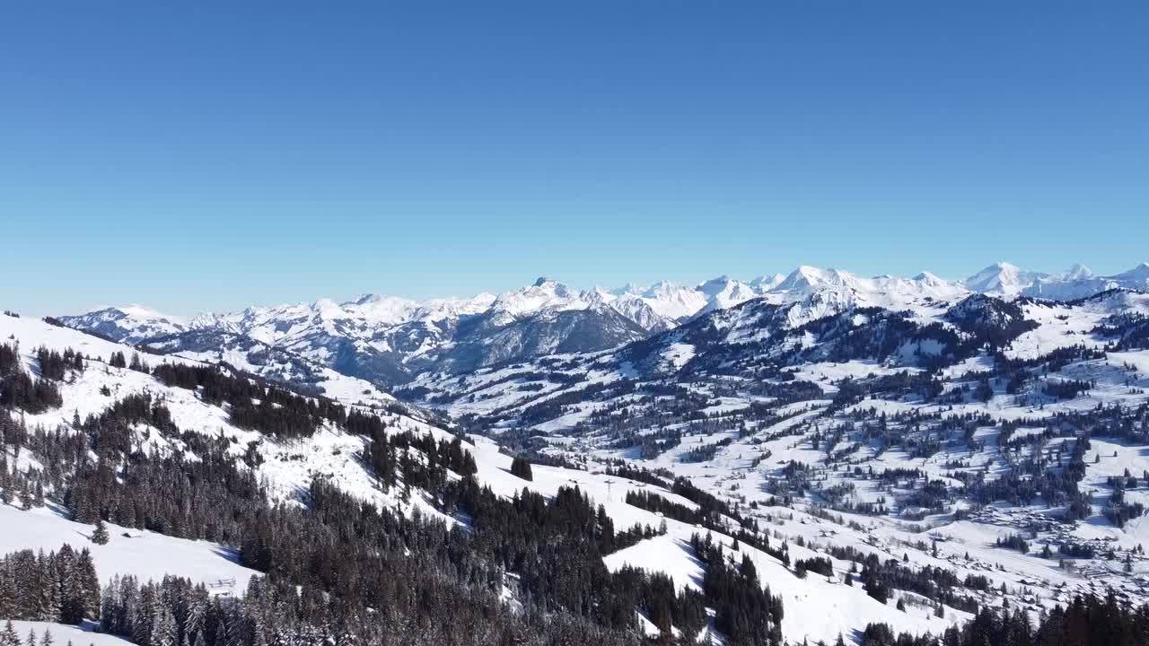 Drohnenflug über eine verschneite schweizer Alpenlandschaft mit Tannen im Vordergrund, bei perfektem sonnigem Wetter mit klarem blauem Himmel