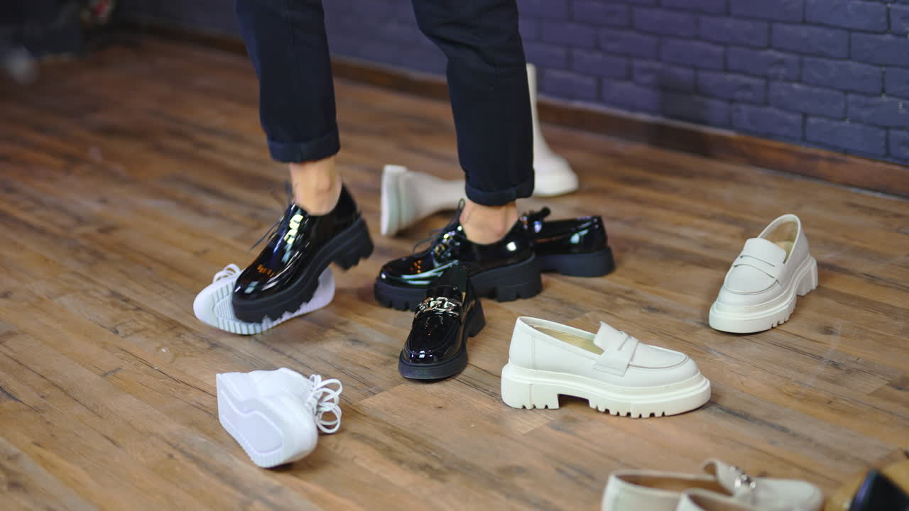 Woman walks in black shoes with tractor soles among other footwear. Black and white footgear scattered on the wooden floor.