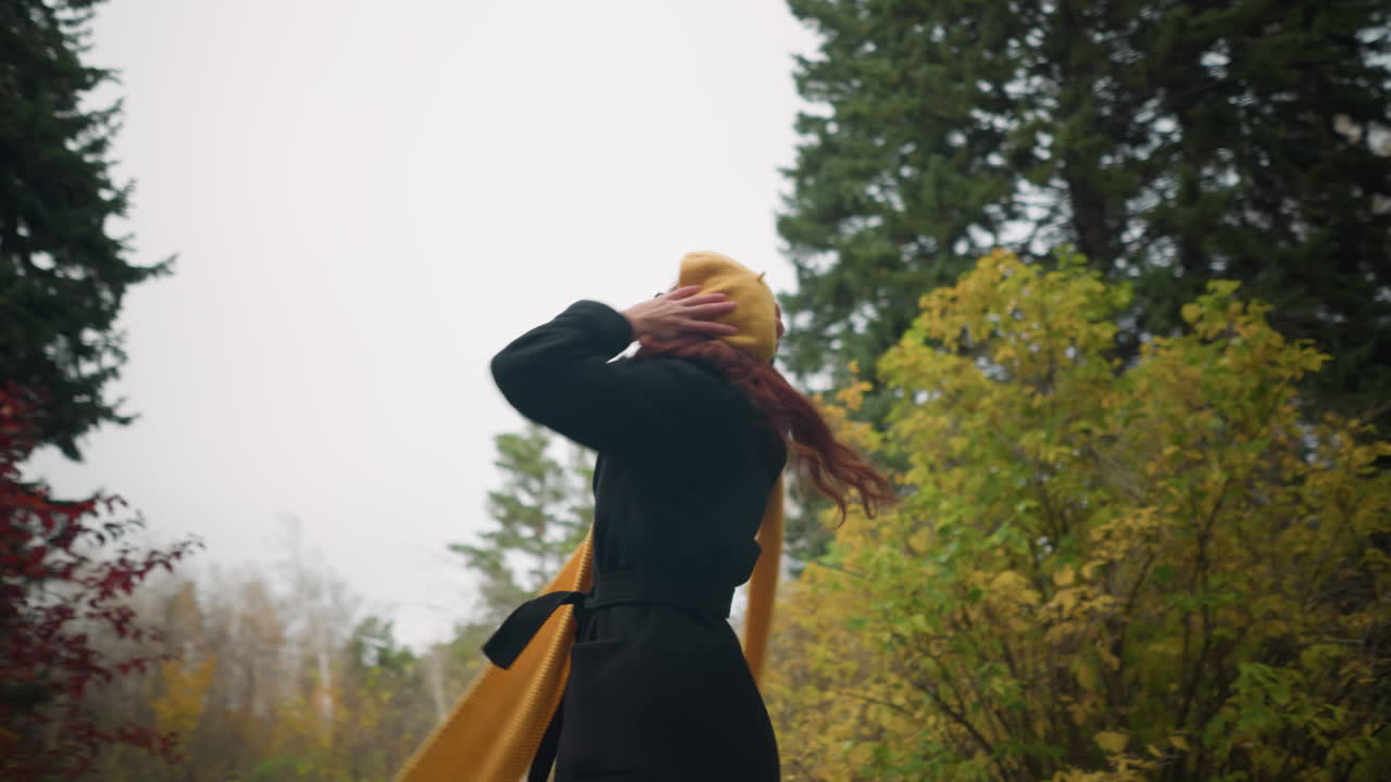 은 곱슬머리를 가진 여자가 기게 돌아서서, 머리에 손을 고, 노란색 베레트와 마플러를 입고, 활기찬 정원 분위기를 즐기면서 흔들고 있습니다.