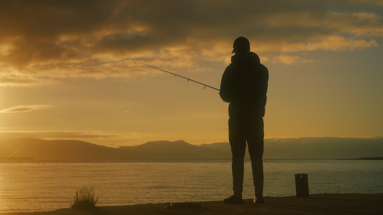Siluet seorang nelayan yang sedang memancing di sepanjang garis pantai Norwegia saat matahari terbenam, dengan cahaya keemasan memantul di air yang tenang dan pegunungan di kejauhan menciptakan pemandangan sinematik