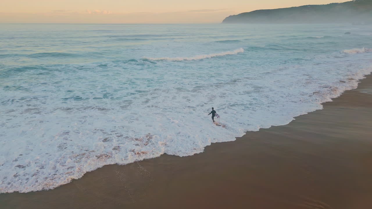 Lonely surfer entering ocean drone shot. Wonderful pastel sky with marine waves