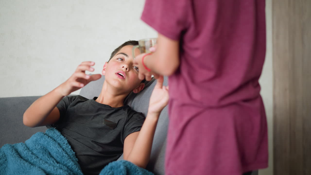 Side view of young girl handing herbal tea to her brother seated on couch, offering care and comfort during illness as he collects and drinks from the cup