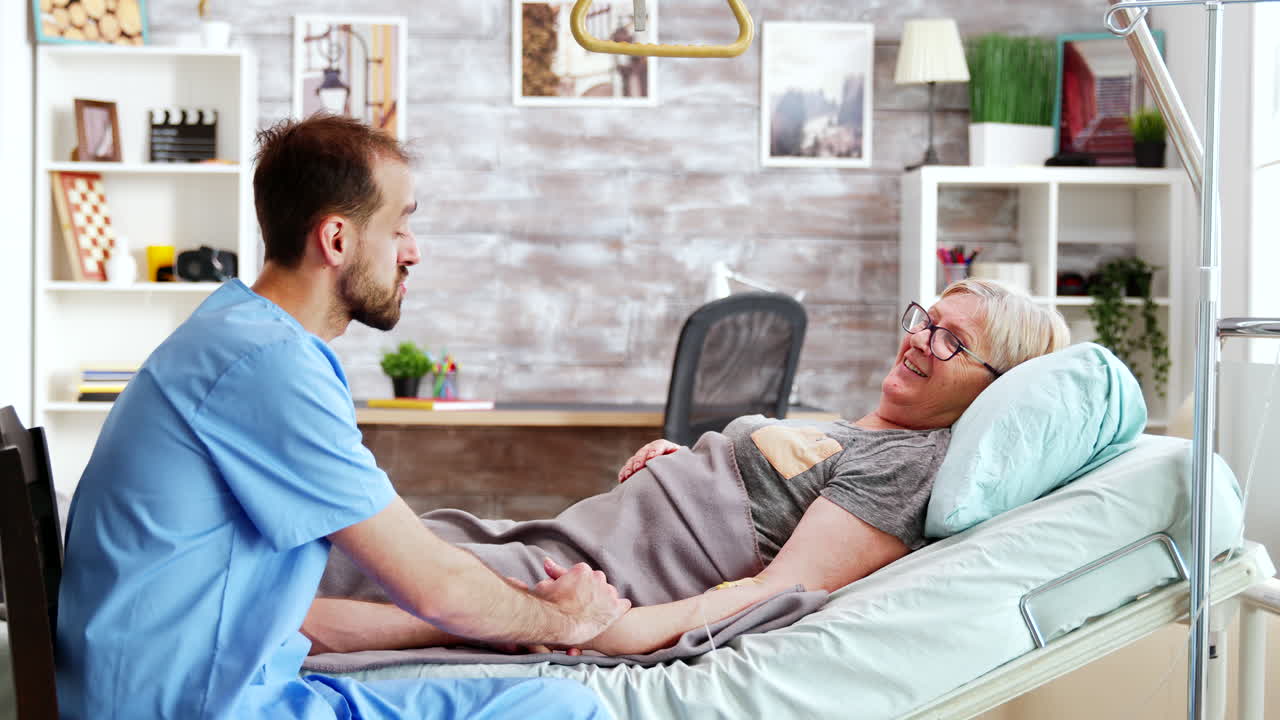 Male caregiver talking with a sick old lady lying in hospital bed