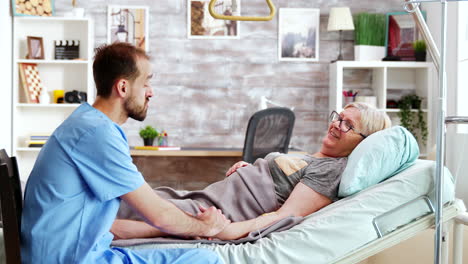 male caregiver talking with a sick old lady lying in hospital bed