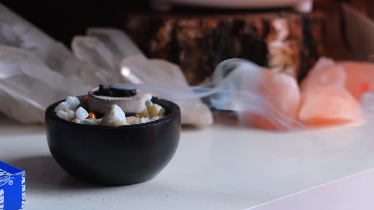 Close up shot of burning incense with white smoke in a bowl with crystal gemstones in the background, in 4K slow motion
