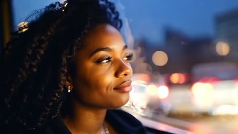 a young woman with curly hair looks out the window of a bus at night. she is sitting in the bus and the city lights are blurred in the background.