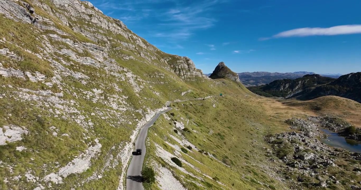 Riprese aeree dall'alto che seguono un'auto su una strada nel Parco Nazionale del Durmitor, Montenegro, mostrando montagne aspre e un ripido strapiombo in una giornata primaverile