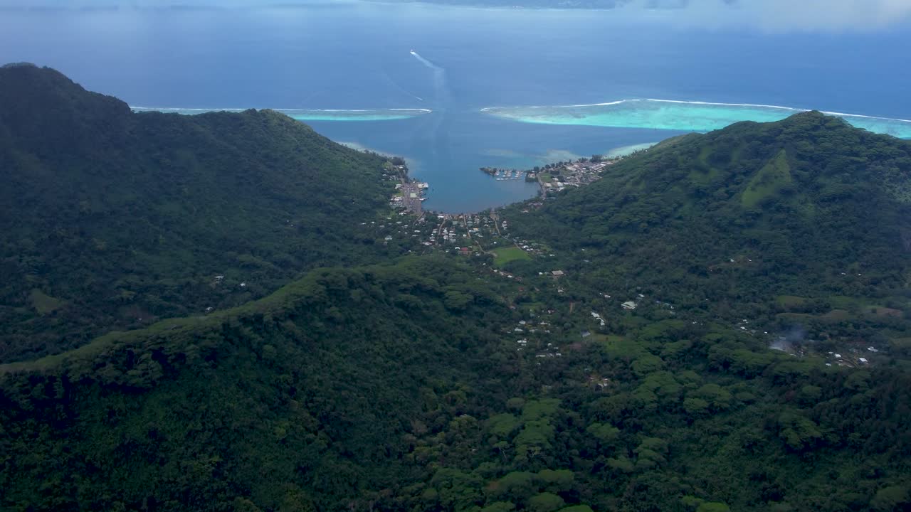 Drone view of mountains a port and a ferry sailing from a green tropical pacific island on a sunny day in Moorea, French Polynesia.