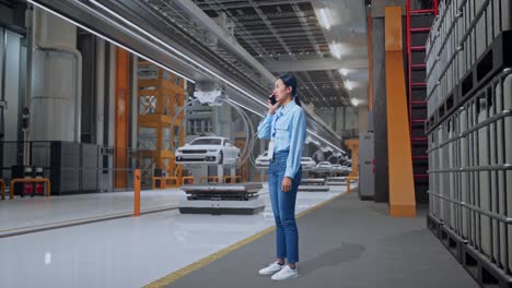 vista lateral completa del cuerpo de una trabajadora profesional asiática de pie en una fábrica de automóviles ev, instalación industrial de alta tecnología, está hablando con alguien en el teléfono