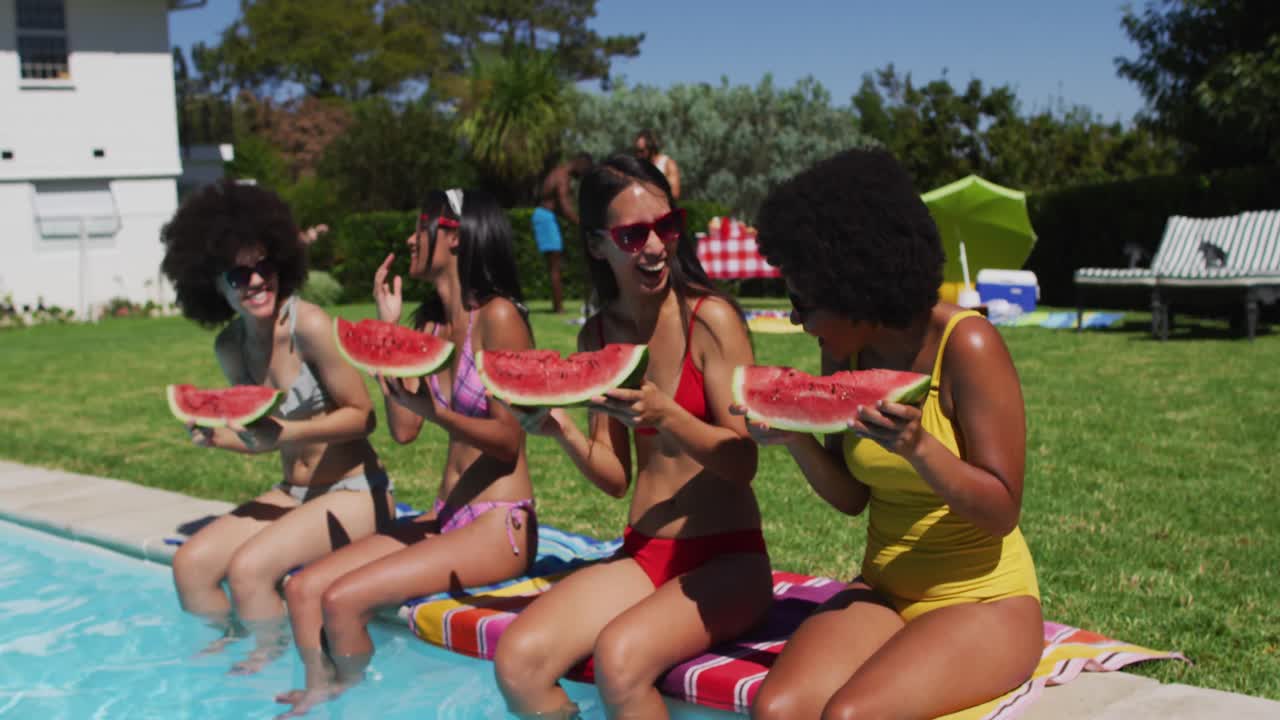 Grupo diversificado de amigas a comer melancia, sentadas à beira da piscina e a conversar
