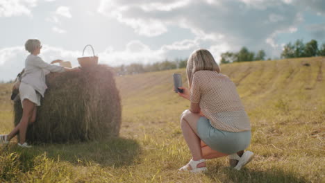 rear view of lady squatting taking photos of her sister in vast farmland with blurred background, warm countryside scene with hay bale and golden sunlight