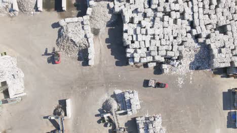 Top-down-drone-shot-of-forklifts-moving-garbage-cubes,-sunny-day-at-a-scrapyard