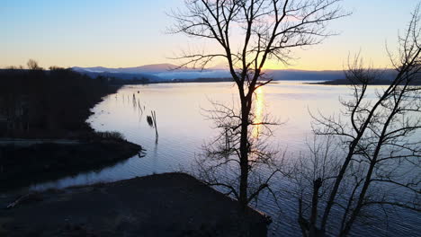 Drone-slowly-panning-to-the-right-behind-trees-with-a-beautiful-sunset-overlooking-a-gorgeous-large-lake