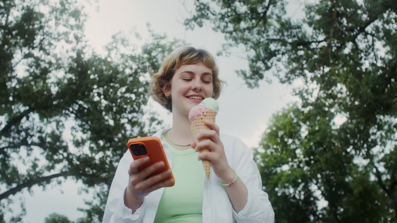 una adolescente disfrutando de helado en el parque mientras usa un teléfono inteligente