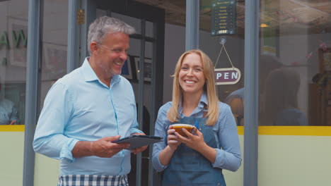 portrait of male and female owners or staff standing outside coffee shop with digital tablet