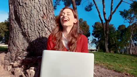 A-happy,-attractive,-young,-caucasian-woman-at-the-park,-typing-on-a-laptop-under-a-tree---gives-a-big-laugh