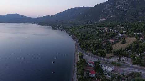 Aerial-view-of-a-coastal-road-in-Greece-with-villages,-forests,-mountains,-calm-sea,-and-boats-on-a-very-early-morning