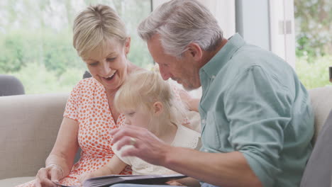 grandparents sitting on sofa with granddaughter at home looking through photo album together