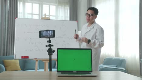 asian male teacher with glasses and green screen laptop shoots video by smartphone camera while teaching chemist at home