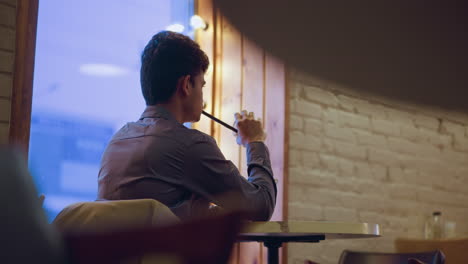 young man sitting in cafe near window with mirror lights, holding paper cup with straw, sipping slowly while staring outside, calm urban indoor mood, relaxed posture, soft evening glow