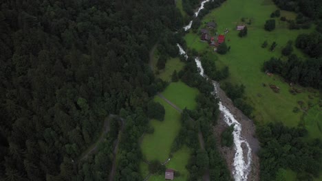 An-aerial-shot-moving-forwards-and-tilting-up,-showing-first-a-beautiful-river-running-down-and-then-revealing-the-cute-and-picturesque-village-in-the-valley-surrounded-by-massive-mountains