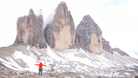 rear view of a woman in an orange hoodie raising her arms after reaching the snowy slope of tre cime di lavaredo