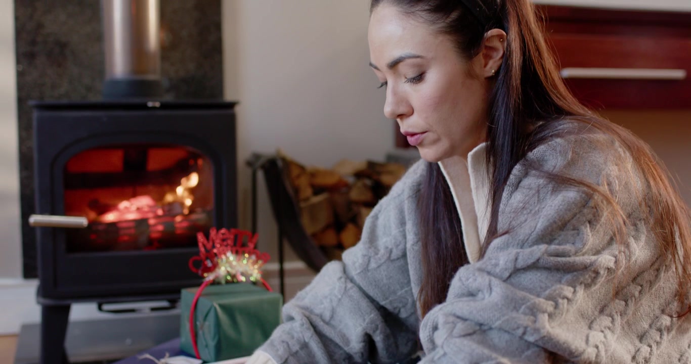 Mujer adulta sentada con las piernas cruzadas en un suelo de madera, preparando regalos navideños y atando un lazo de cinta dorada.