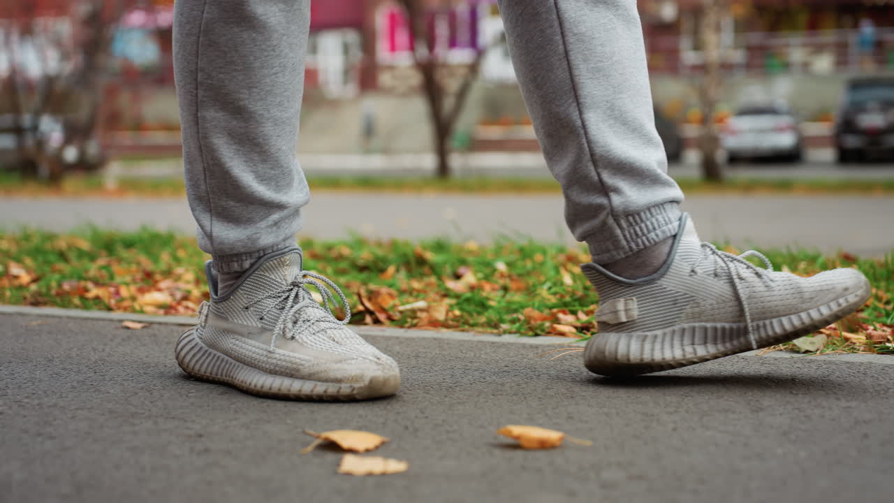 Leg view of sports person walking along tarred path with autumn leaves scattered across ground, car moving and others parked near road, building and trees visible across in urban park setting