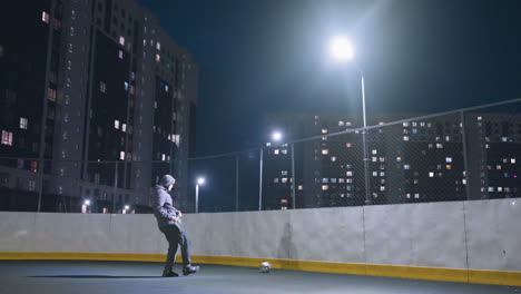 vista trasera de un atleta con capucha pateando una pelota de fútbol contra la pared bajo la iluminación brillante del estadio por la noche, la pelota refleja la silueta en la pared con el paisaje urbano y los edificios de apartamentos iluminados