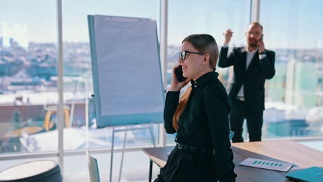 business people talking on their phones indoors. smiling woman having a conversation through the phone in modern office. businessman near the window with city background.