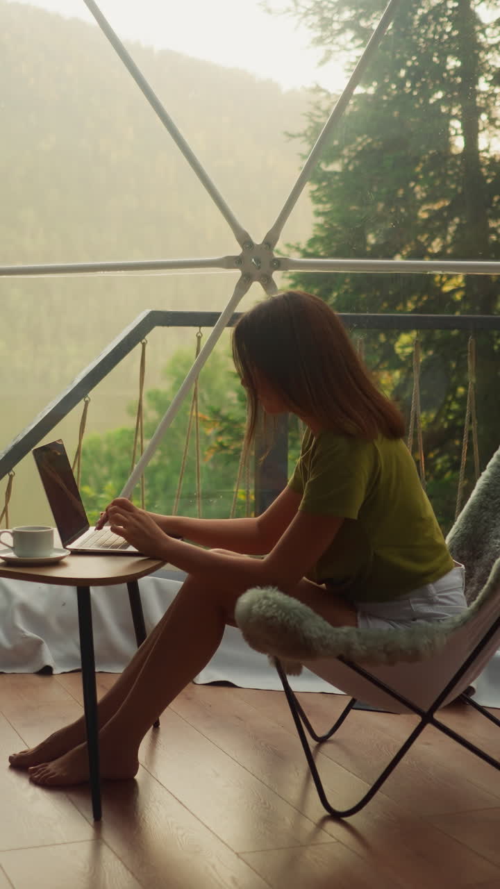 Woman works on laptop on vacation in bright room of glamping house. Young woman alone sits in armchair and types text on keyboard with cup of coffee on table