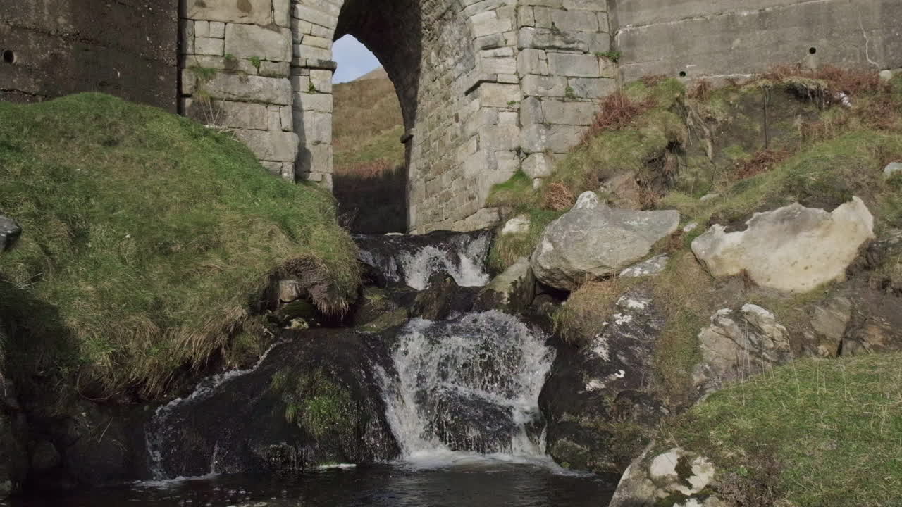 Medium wide shot of small water fall running under a stone bridge. Grass on each side of the stream. Handheld camera shot with slow motion.