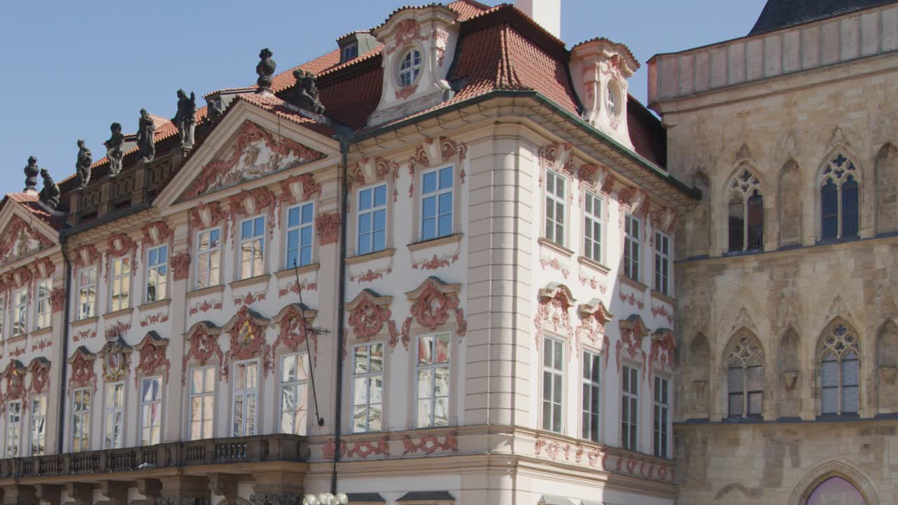 Camera slowly pans across ornate historic buildings in Prague city square under clear daylight, highlighting architectural details and vibrant facades with steady movement