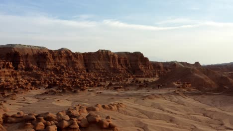 Gorgeous-descending-aerial-drone-shot-of-the-beautiful-Goblin-Valley-Utah-State-Park-with-small-strange-mushroom-rock-formations-below-and-large-red-and-white-Butte's-in-the-background