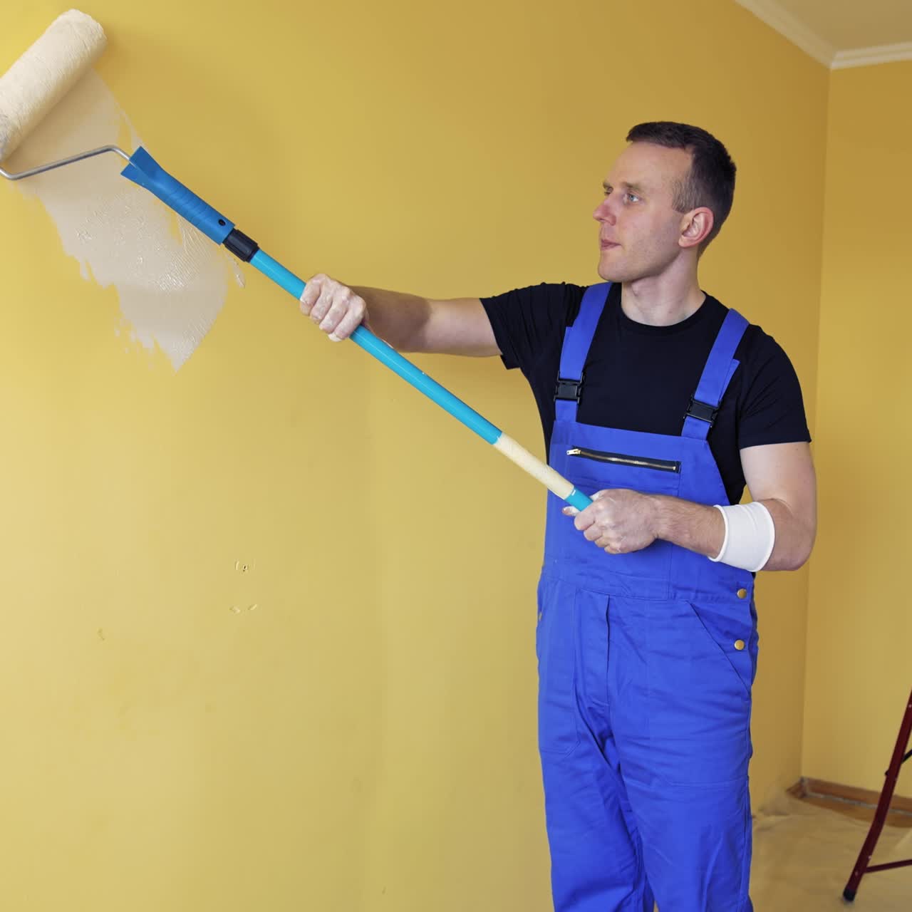 Room renovation at home. Builder specialist painting yellow wall into new color. Portrait of a young craftsman in overalls starts painting wall with a paint roller