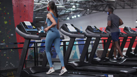 fit caucasian woman and african american man wearing face masks running on treadmill in the gym