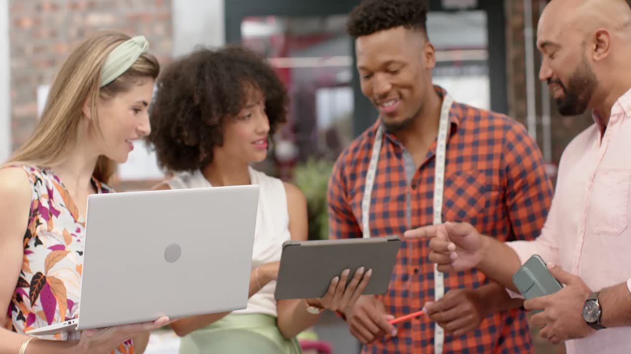 태블릿과 노트북을 사용하는 다양한 행복한 디자이너들이 패션 스튜디오에서 느린 동작으로 토론하고 있습니다.