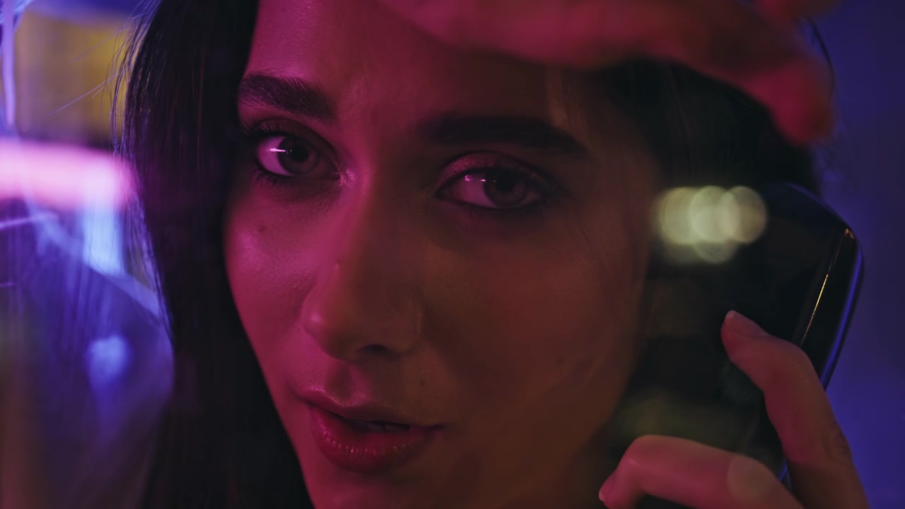 Primo piano del volto di una giovane donna che parla al ricevitore di un telefono pubblico, luce al neon, strada cittadina di notte
