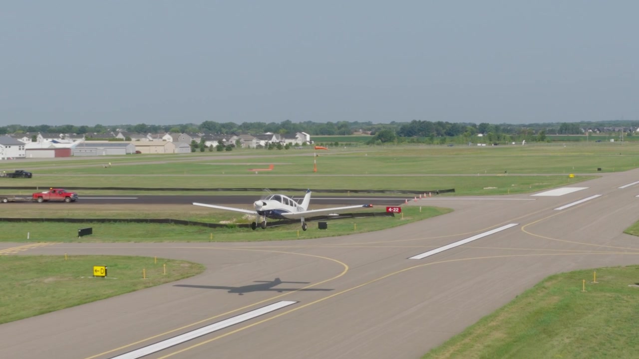 Ripresa aerea in movimento di un piccolo aereo che decolla dalla pista in un aeroporto regionale con hangar sullo sfondo.