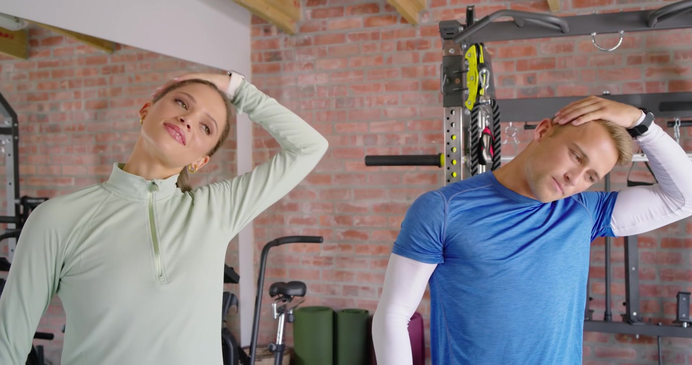 Diverse couple warming in sportswear after smile, holding arms, tilting necks in home gym near rack