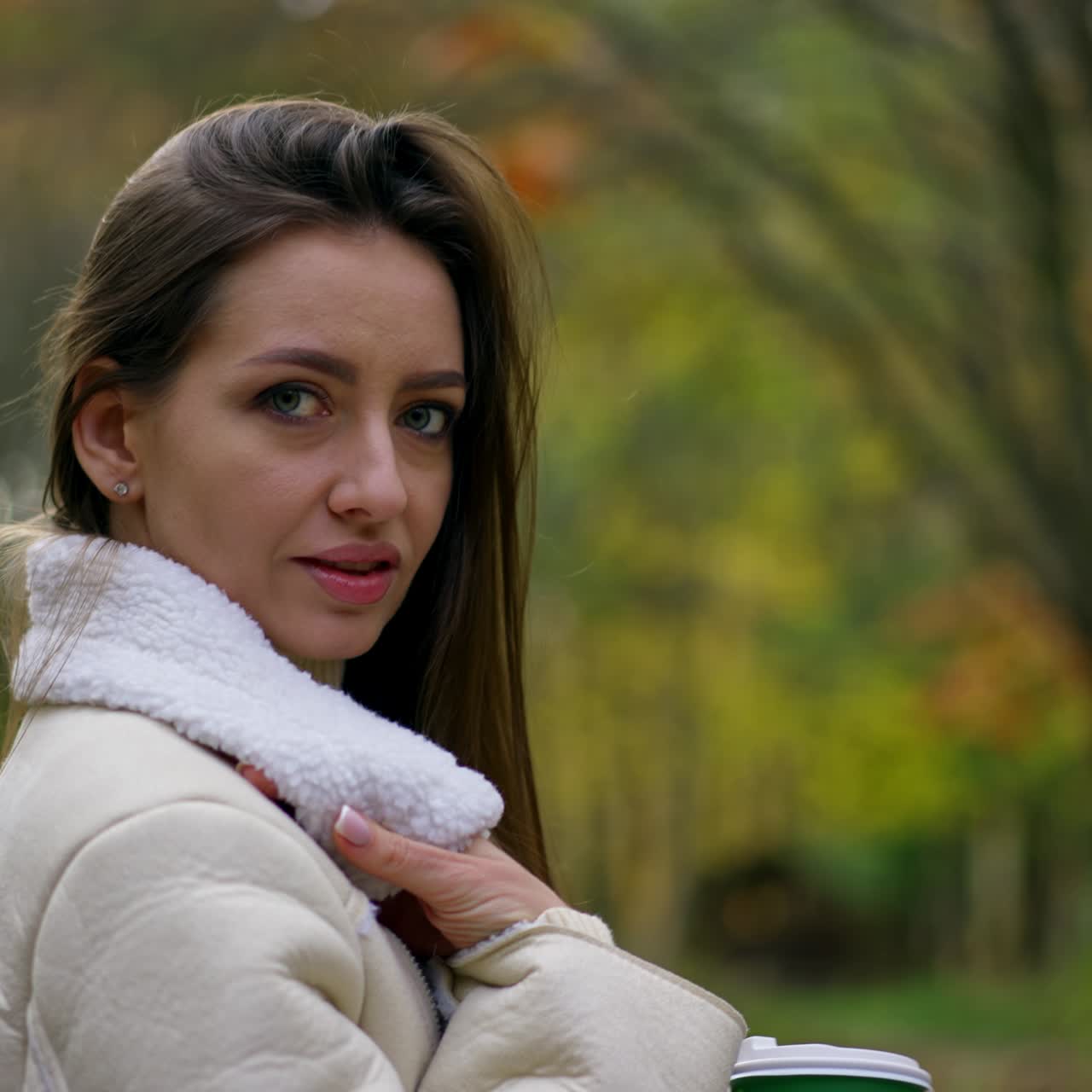 Beautiful big-eyed brunette in white jacket stands in the park her side to camera looking at it. Lady looks at the paper cup in her hands touching her hair