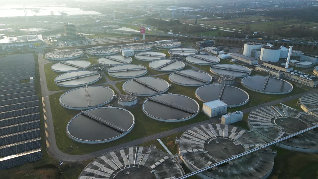 High quality aerial drone footage showing circular wastewater clarifier basins and adjacent solar panel installations at the Harnaschpolder treatment facility in the Netherlands during sunset