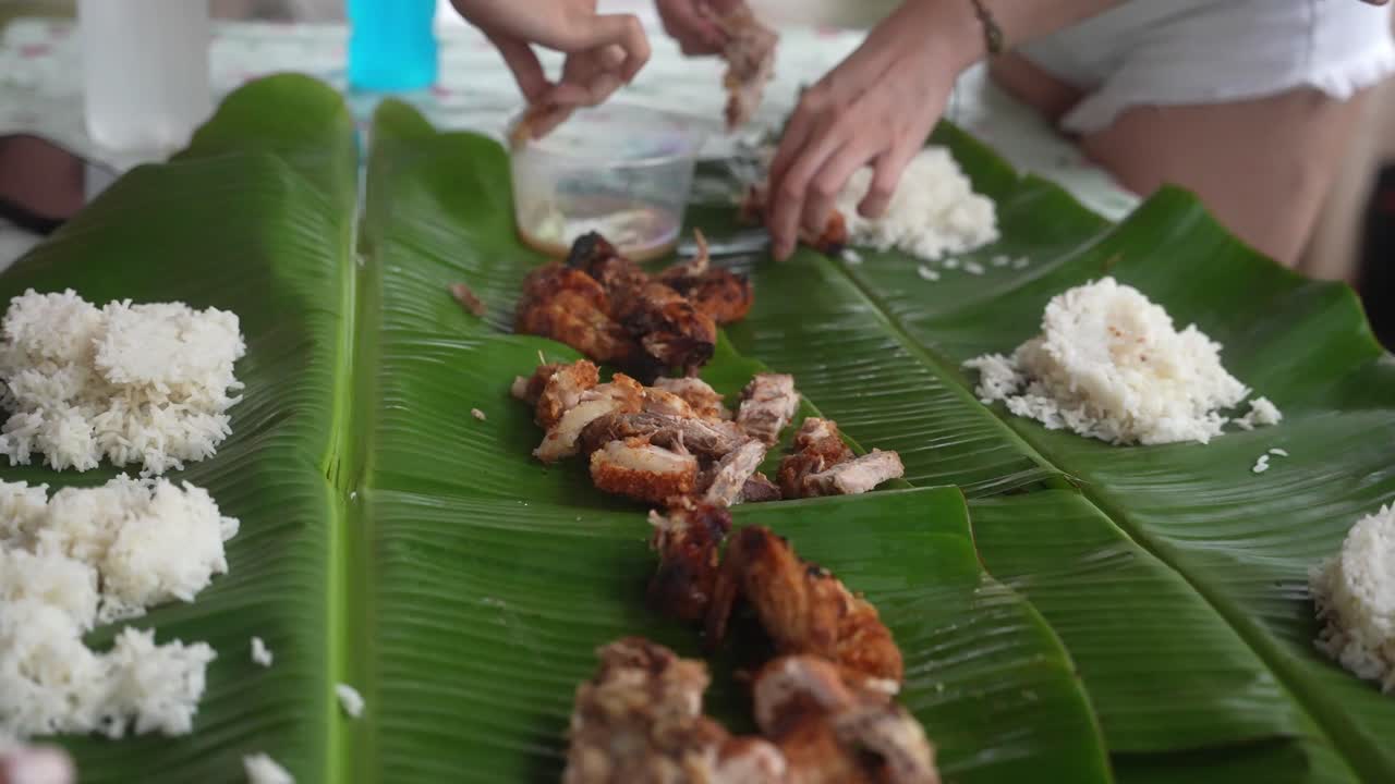 Handheld medium shot captures a traditional Philippine boodle fight with rice and grilled meat on banana leaves, as hands reach in for a shared, hands-on meal