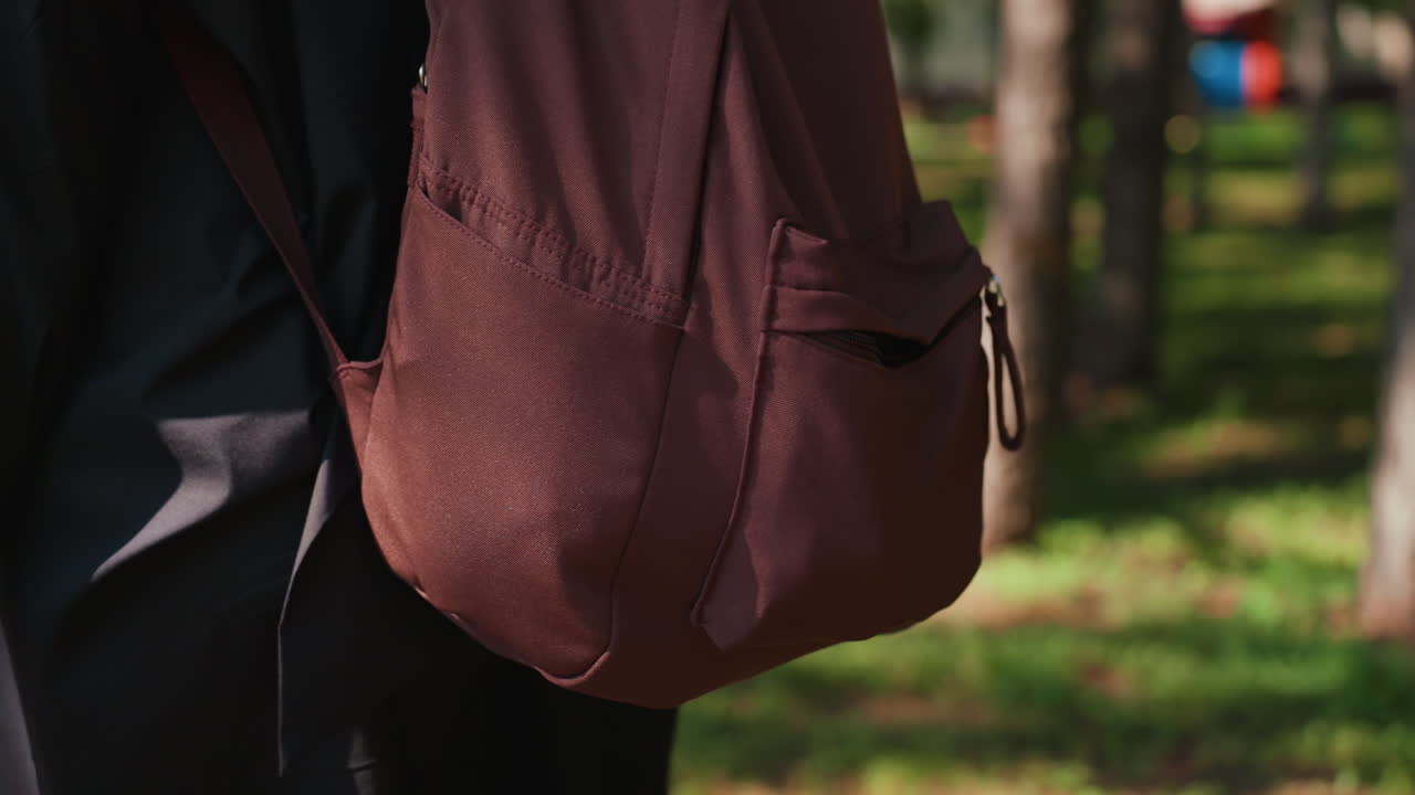 가방을 메고 공원을 하이킹하는 사람, 낮 동안 그늘진 공원 산책로를 이동하는 캐주얼한 개인, 햇빛 아래 나무가 늘어선 공원 길을 꾸준히 걷는 학생 또는 여행자