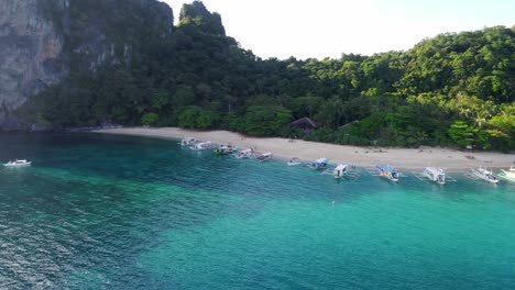 Aerial,-island-Hopping-Boats-lined-up-at-white-sandy-beach-of-Helicopter-Island,-El-Nido